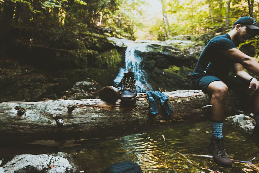 man sitting on tree log beside boots, man sitting on tree trunk lying on river at daytime HD wallpaper