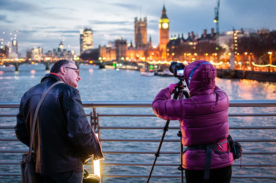 two people standing in front of silver fence, man standing beside person taking a picture of body of wate r HD wallpaper