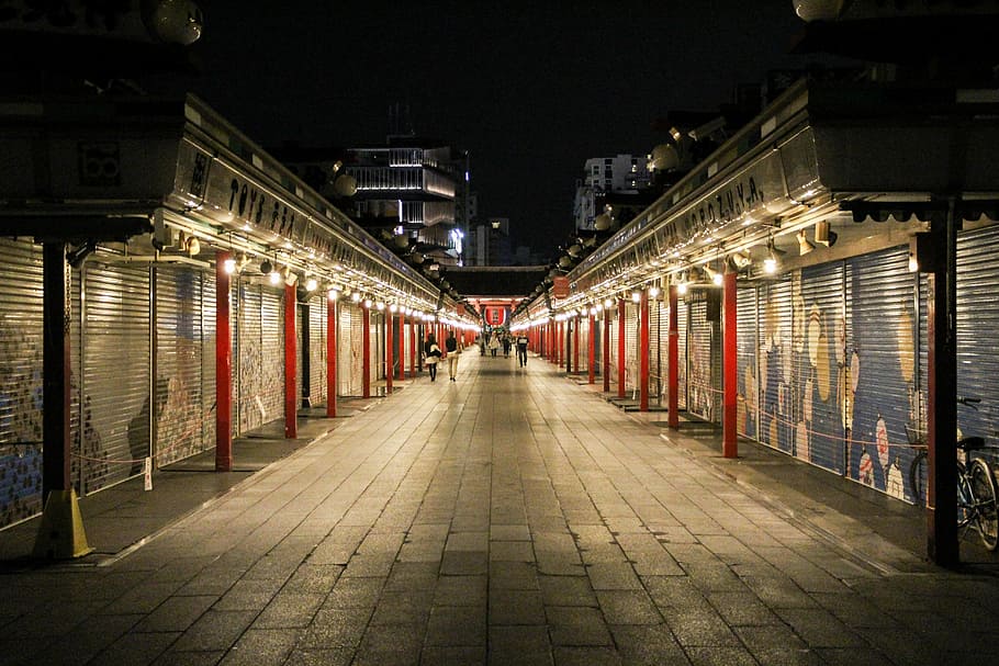 empty pathway beside stores with roll top doors at night time, people walking in the city during nightime