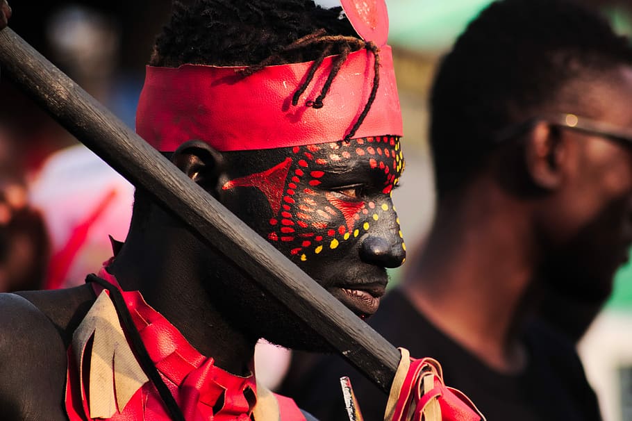man with red and yellow face paint, man wearing red and beige top holding rod