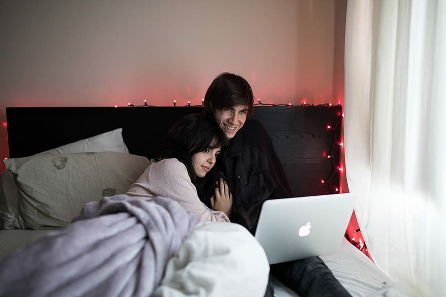 couple on bed using MacBook Air beside of window with white curtain, couple sitting on bed beside covered window
