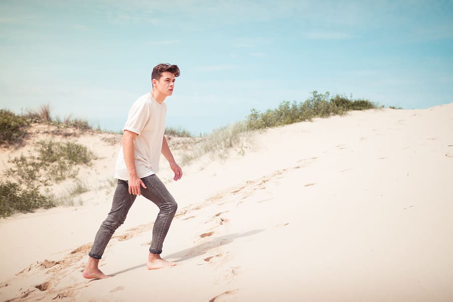 man wearing white t-shirt standing on sand field during daytime, man walking with barefoot