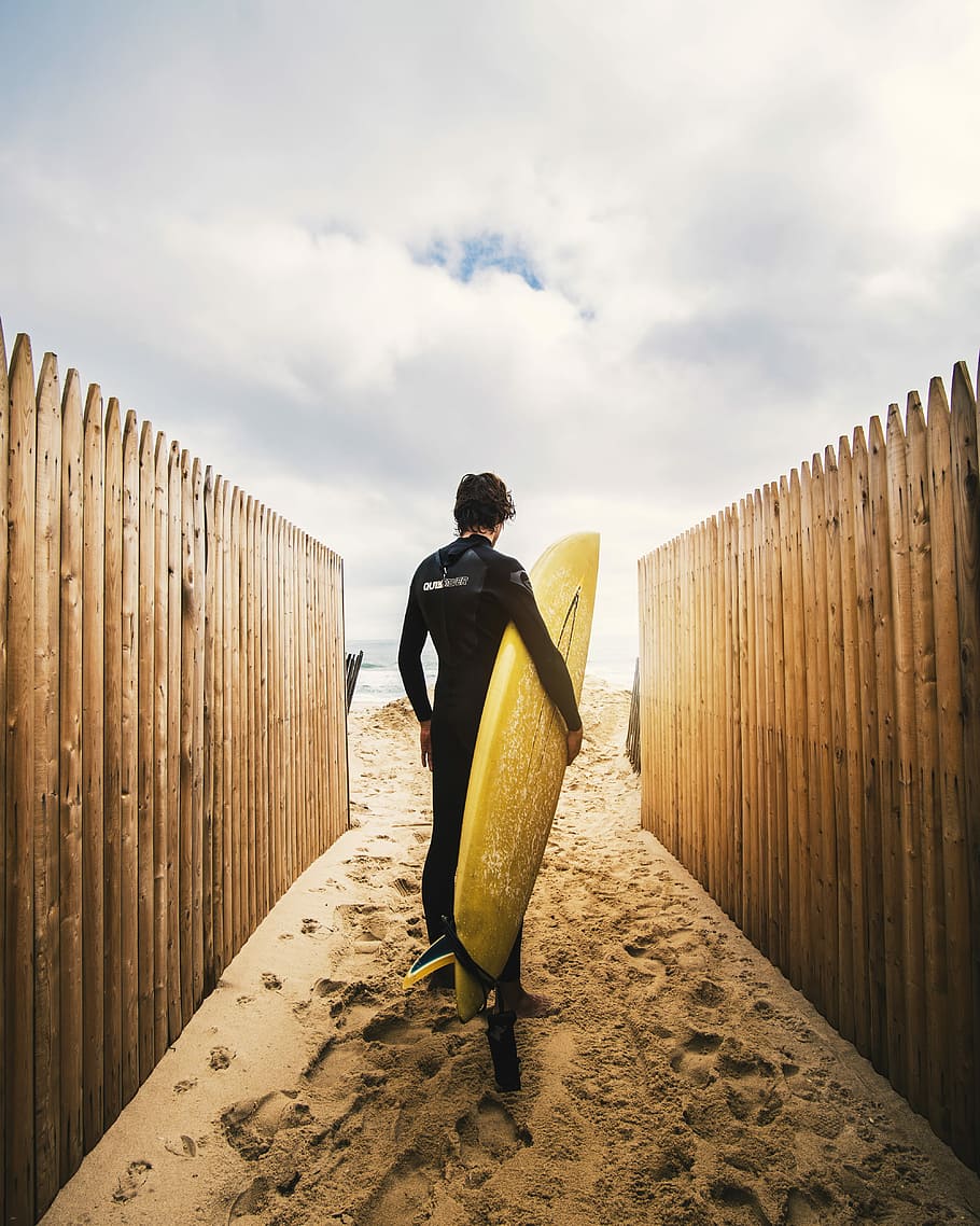 person standing between privacy fences while holding yellow surfboard during daytime, man holding surboard HD wallpaper