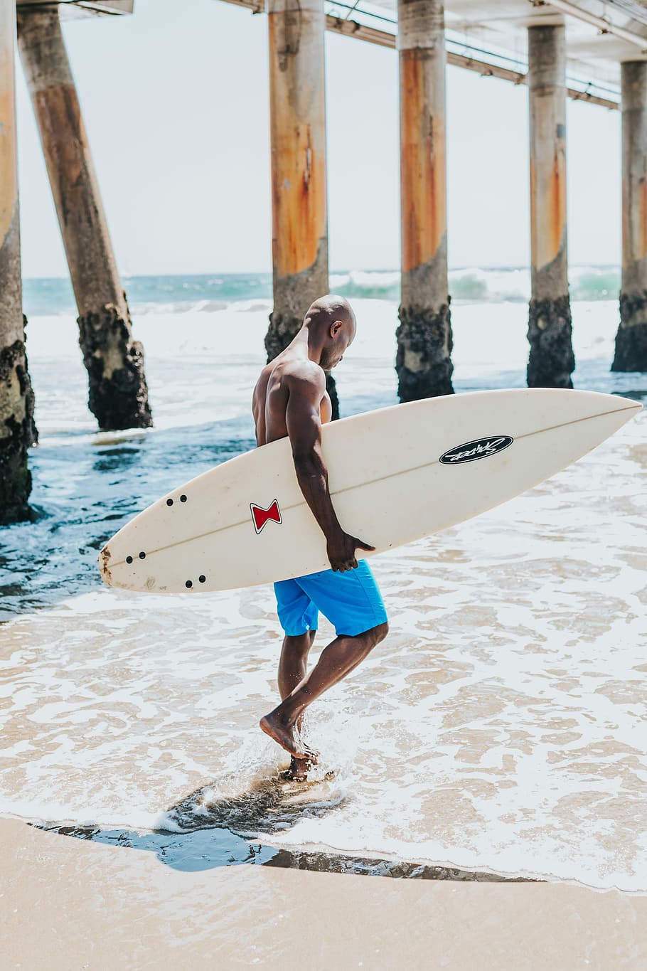 man holding white surfboard walking across ocean, man walking on shore carrying surfboard during day