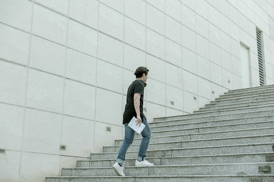 man holding white blanket paper walking on gray stair, man carrying white book while walking upstairs beside white concrete wall