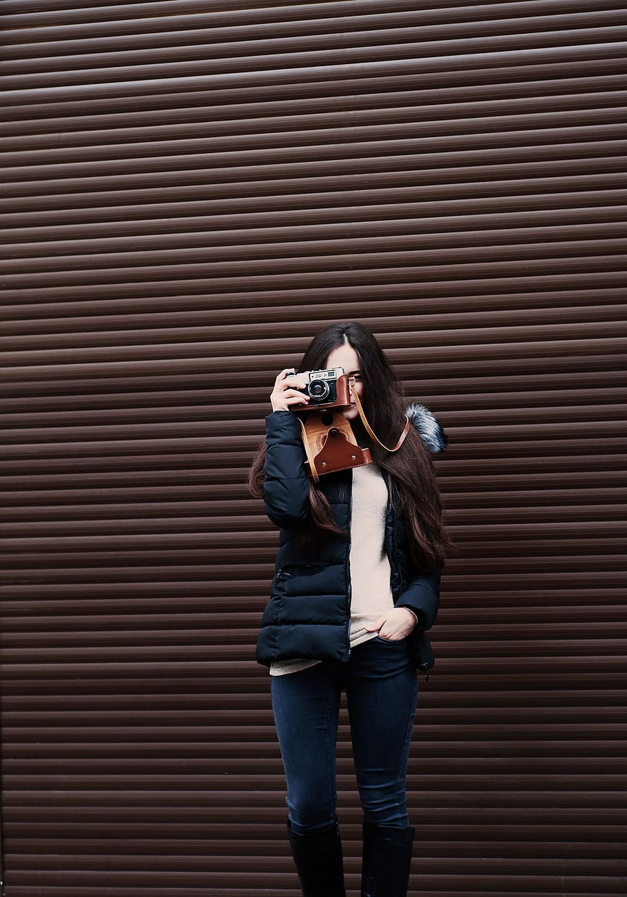 woman standing holding camera taking photo, woman holding black camera standing beside brown shutter door HD wallpaper
