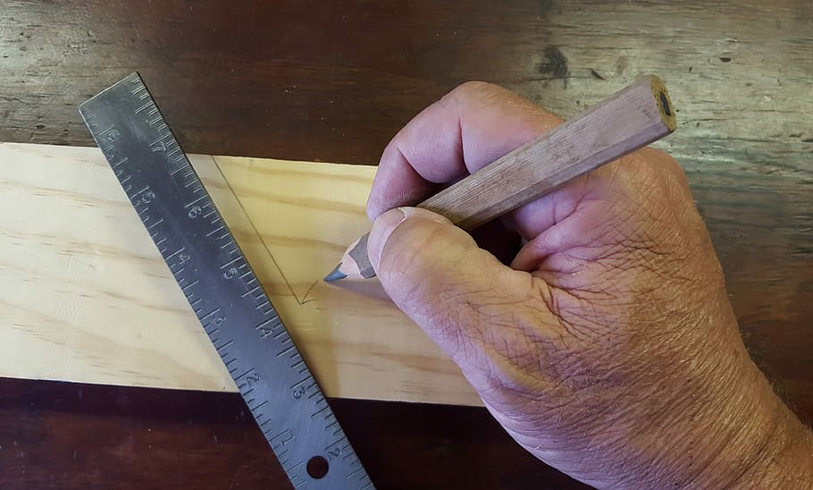 person holding brown wooden pencil beside stainless steel ruler while measuring brown wooden slab