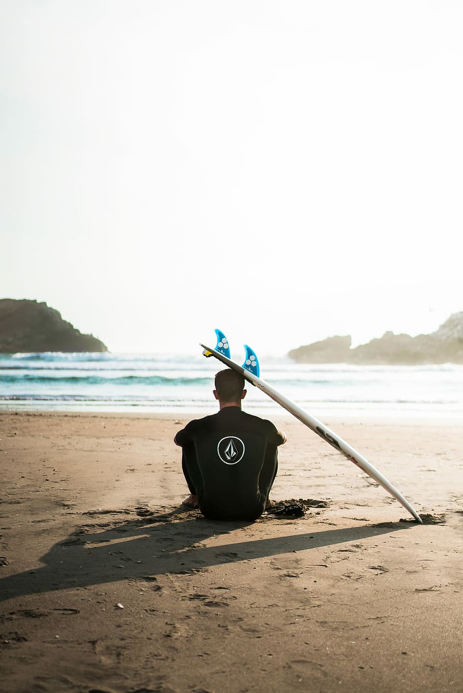 man sitting on sand beside surfboard facing sea during daytime, man sitting on sea shore beside surfboard HD wallpaper