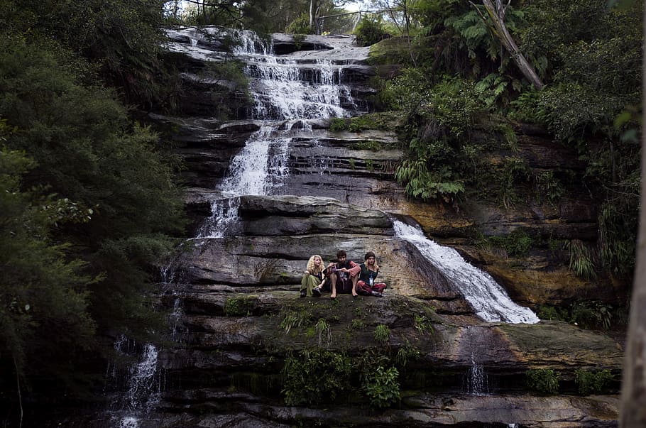 three person sitting on rock formation in waterfalls surrounded with trees at daytime, three person on cliff of waterfalls HD wallpaper