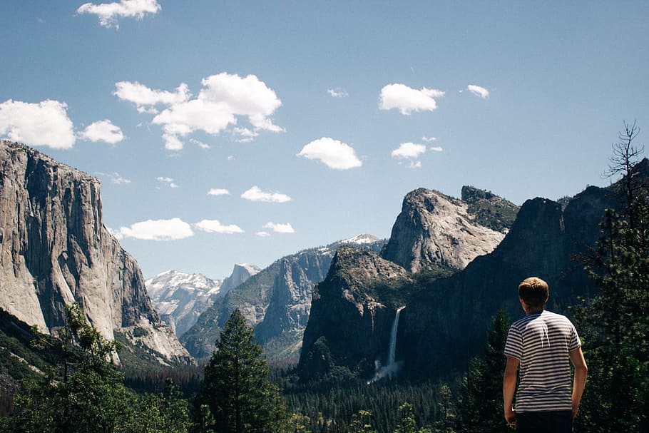 man standing next to mountains with waterfalls and trees, man staring at the mountain range during daytime HD wallpaper