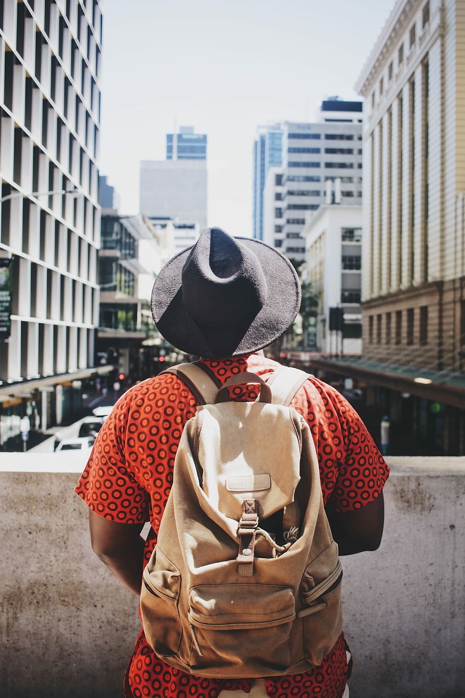 person wearing black hat standing beside concrete rail during daytime, man wearing backpack and facing the high rise building HD wallpaper