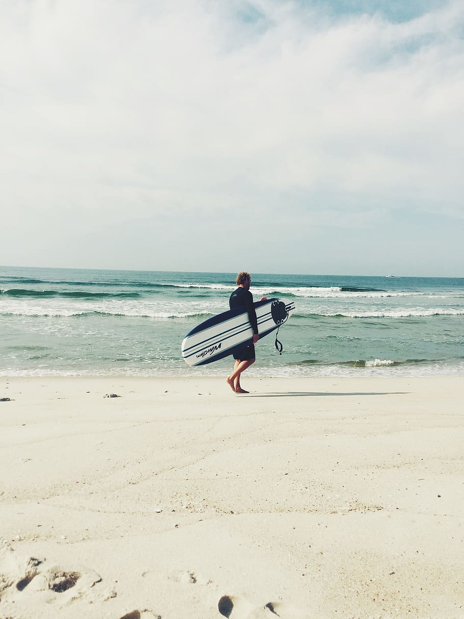 man carrying white and blue surfboard walking on the seashore HD wallpaper