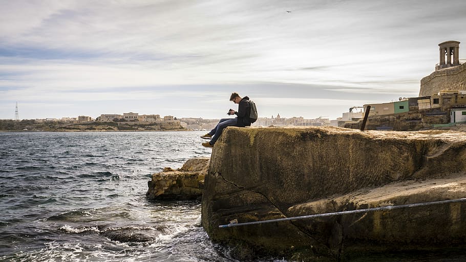 man sitting on brown rock near sea at daytime, man sitting on edge holding camera HD wallpaper