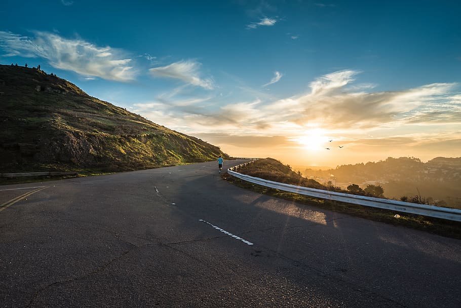 person standing beside road guard rail near green mountain at golden hour HD wallpaper