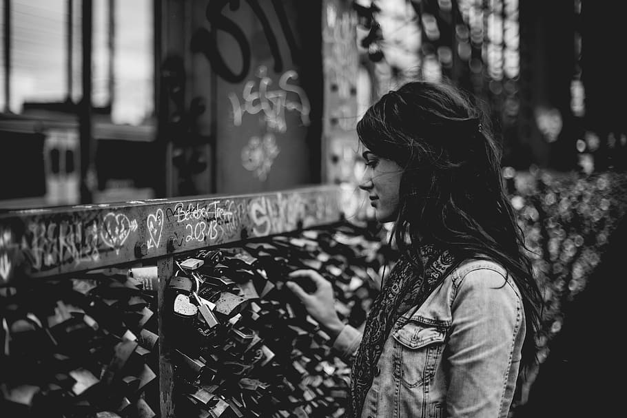 woman holding padlock on bridge, grayscale photography of standing woman front of brown wooden fence