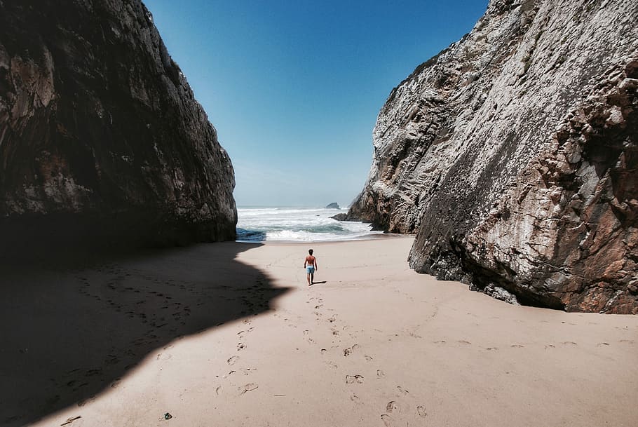 person standing on beige sand during daytime, man walking toward sea on brown sand between rocks at daytime