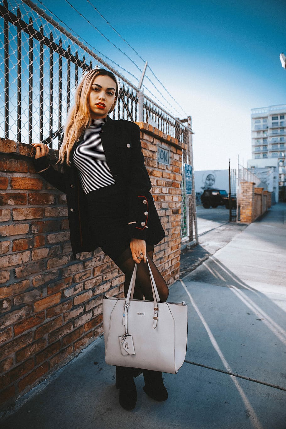 woman wearing black coat standing while holding tote bag besides concrete fence during daytime, woman carrying tote bag while leaning on brick wall