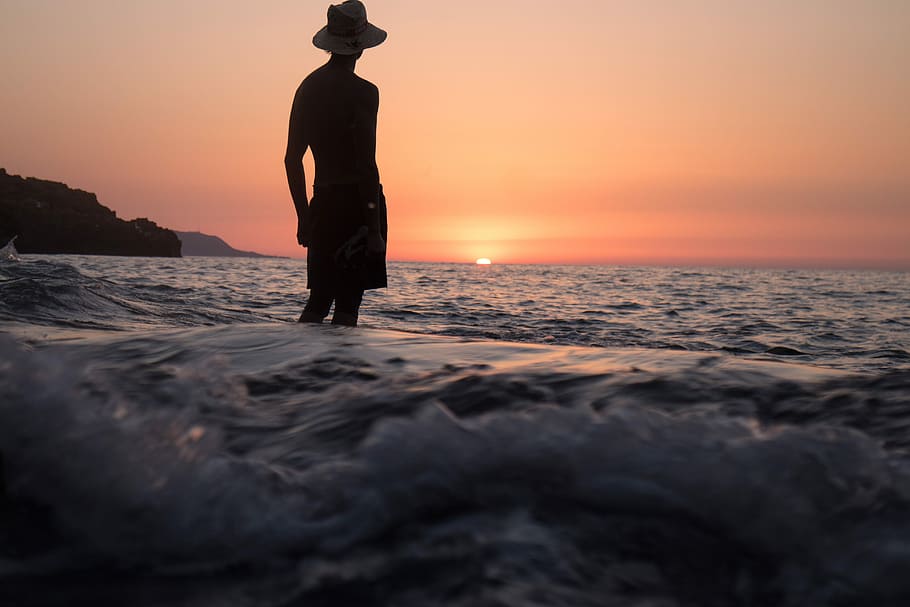 silhouette of person standing on shallow seashore facing sunset, silhouette of man with hat near ocean HD wallpaper