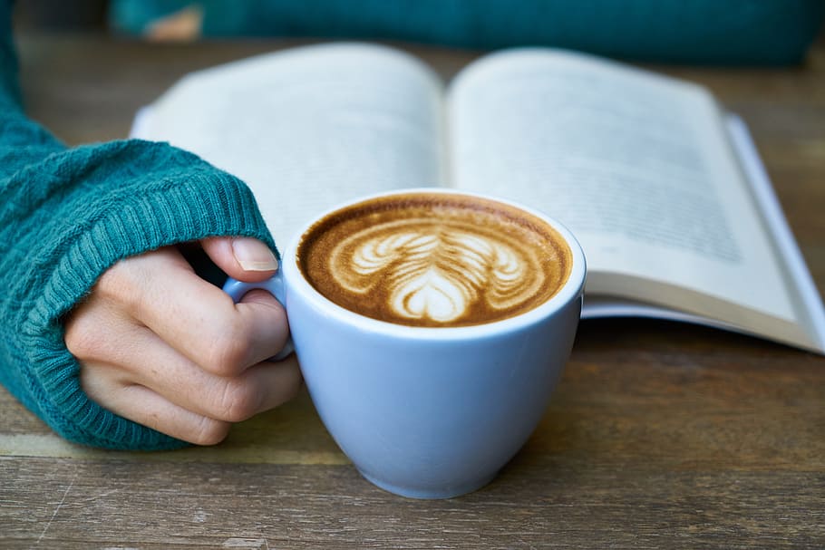 person holding blue ceramic cup with coffee inside and book behind
