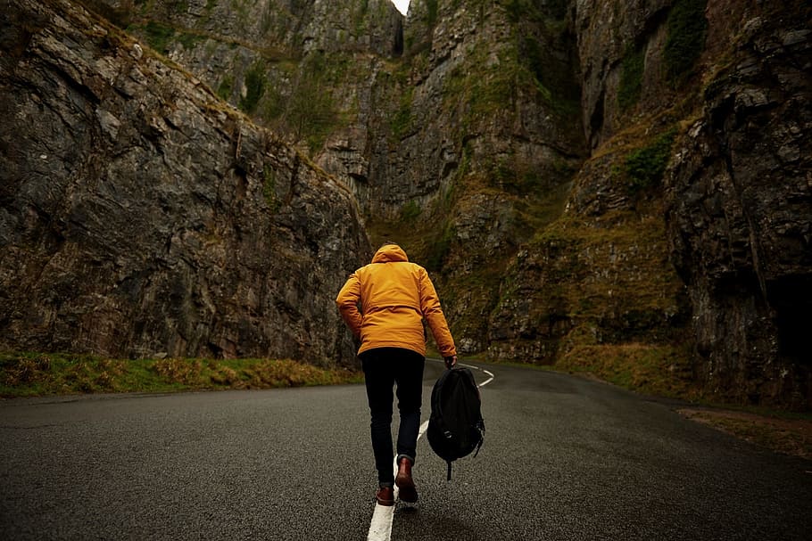 photo of person wearing yellow hoodie and black pants holding backpack walking on roadway between mountain HD wallpaper