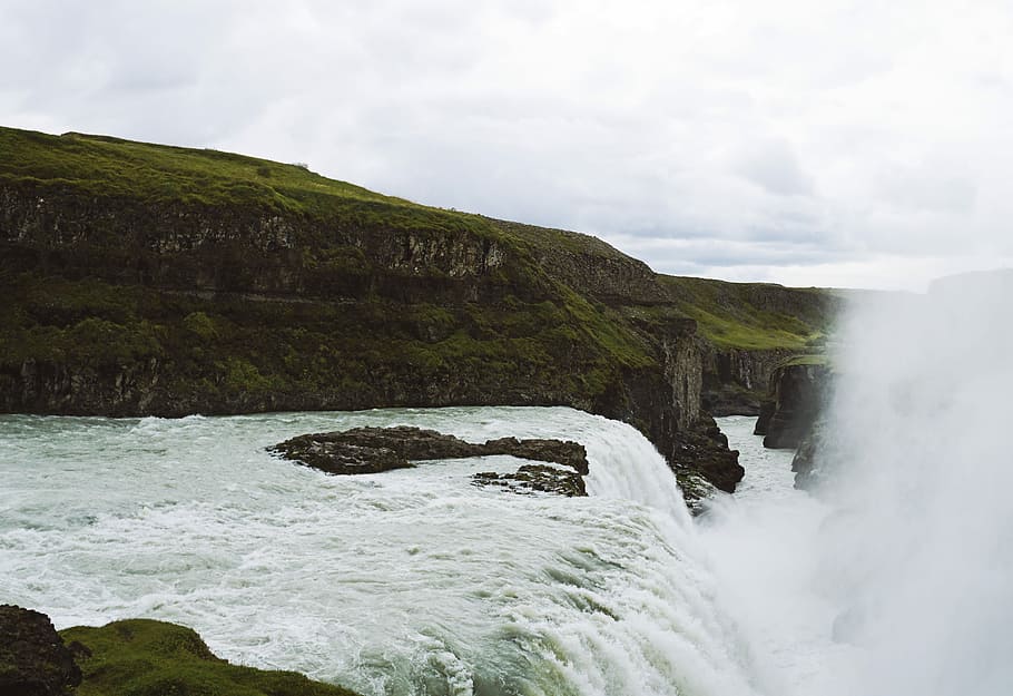 landscape photo of river near green grass covered hill under cloudy sky, waterfalls near mountains at daytime HD wallpaper