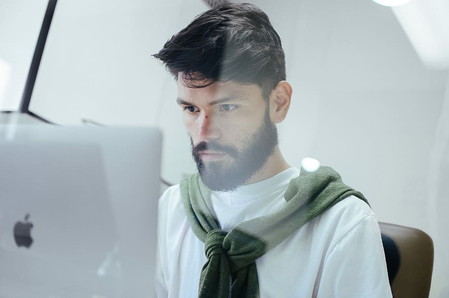 man facing MacBook Pro, man wearing white crew-neck top, beard HD wallpaper