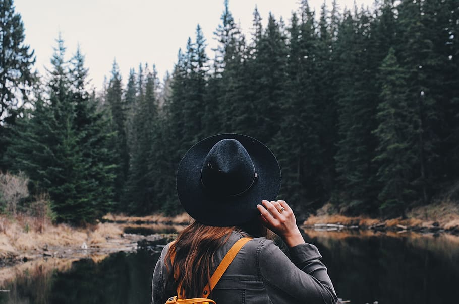 woman holding her hat while standing in front of river beside forests, woman holding her hat looking at trees