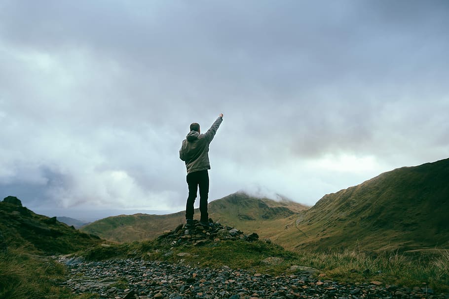 man standing on rocks near mountains pointing sky during daytime, man raising his hand stepping on gray stones, HD wallpaper