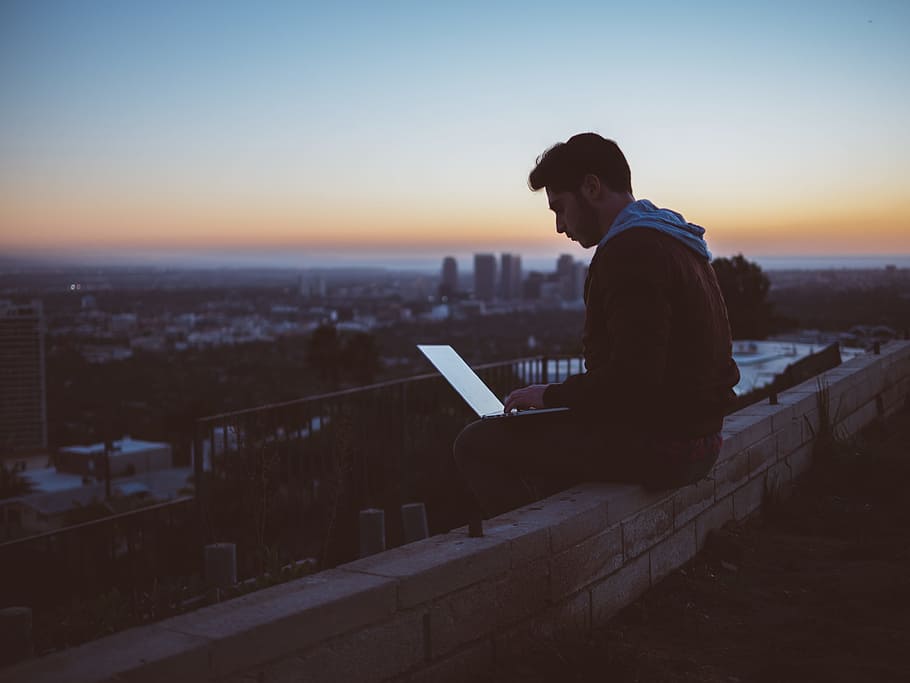 man sitting on concrete brick with opened laptop on his lap, man sitting on edge of building while using laptop computer HD wallpaper