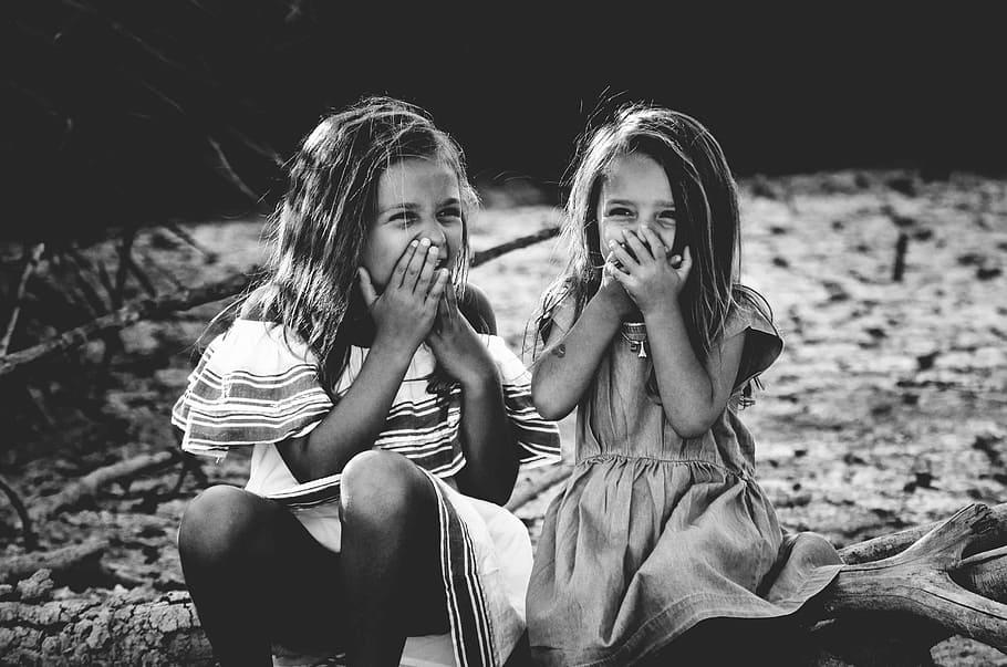 grayscale photography of two girls closing their mouths, two girl's sitting on floor and holding there mouth while laughing