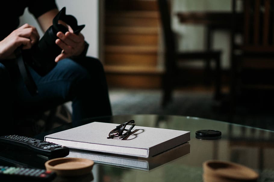 eyeglasses on top of white book, person holding DSLR camera in front of glass-top table HD wallpaper