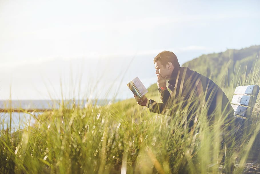 man wearing blue jacket sitting on bench reading books facing body of water HD wallpaper