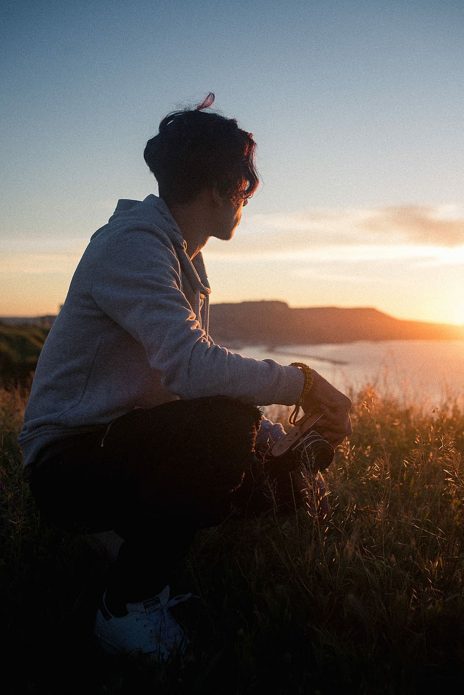 man sitting on rock while holding camera, man sitting on green grass holding camera while watching sunset HD wallpaper