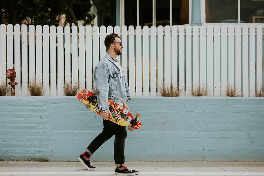 man carrying long board walking beside white fence, man in blue denim button-up jacket and black pants holding multicolored longboard during daytime