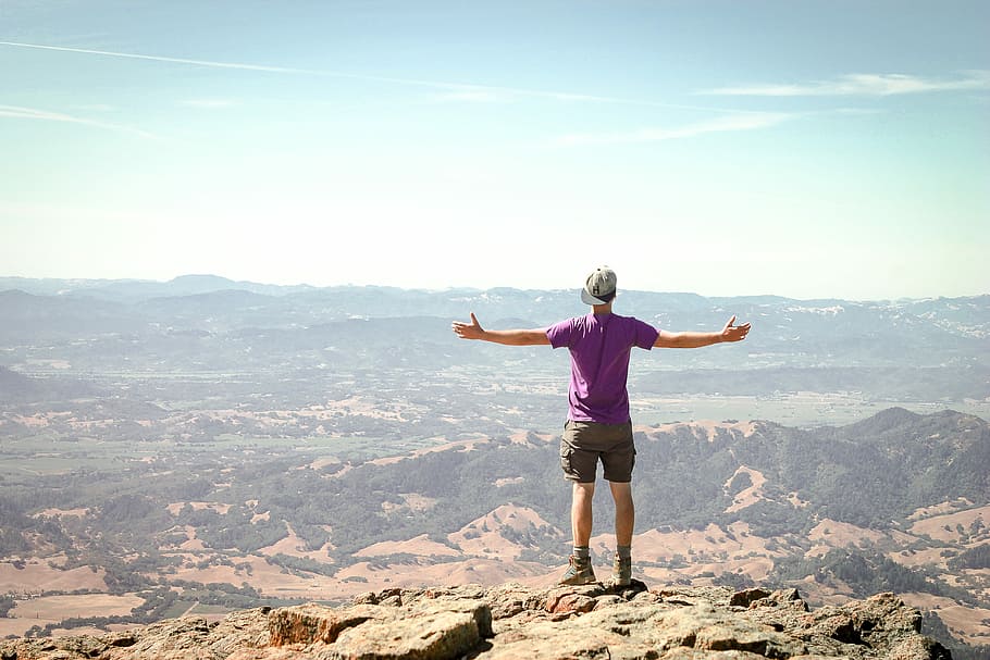 man stand on brown mountain under blue sky, photography of man standing on rock mountain HD wallpaper