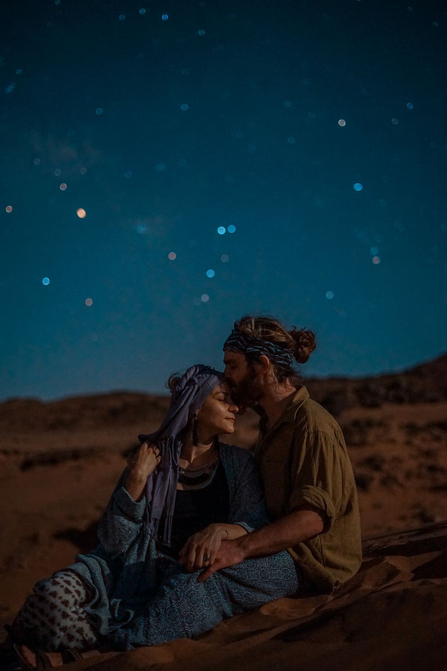 man and woman sitting on desert sand under blue sky during nighttime, man kissing woman's forehead