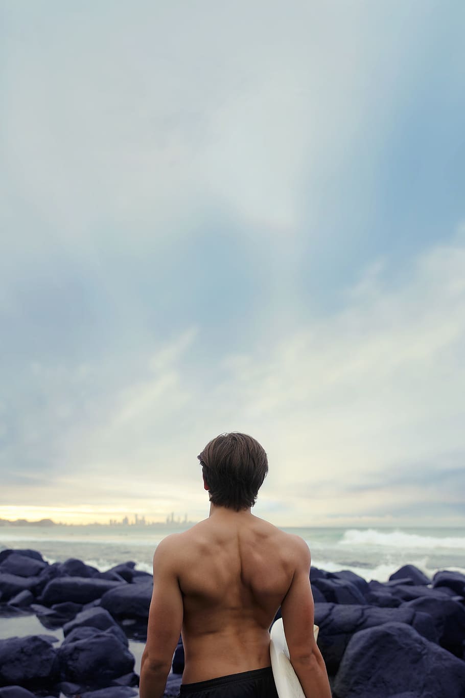 person standing carrying surfboard near seashore, man standing on rocky beach shore holding white surfboard HD wallpaper
