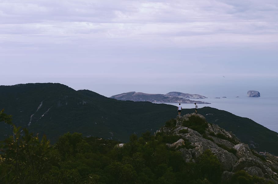 two men standing at top of mountain near sea, two men on peak of stone mountain near body of water HD wallpaper