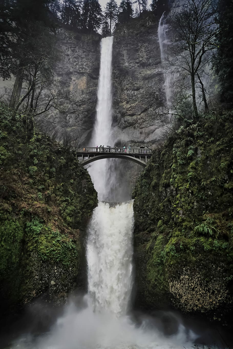 waterfalls in mountain, people passing on grey arch bridge near waterfall