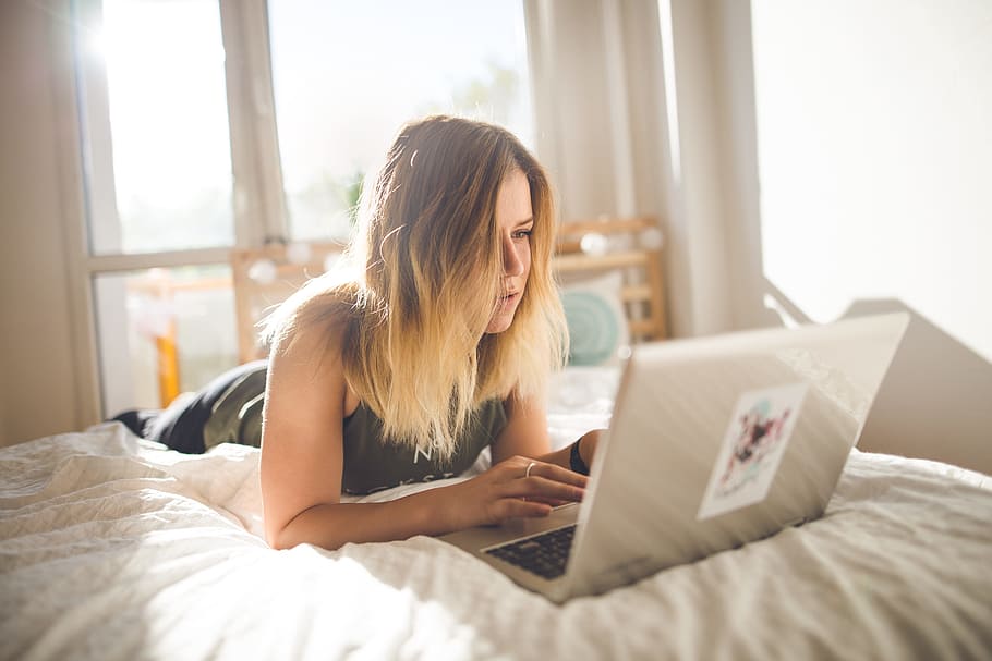woman in black tank top in front of silver laptop computer at daytime, woman on bed while using gray laptop computer