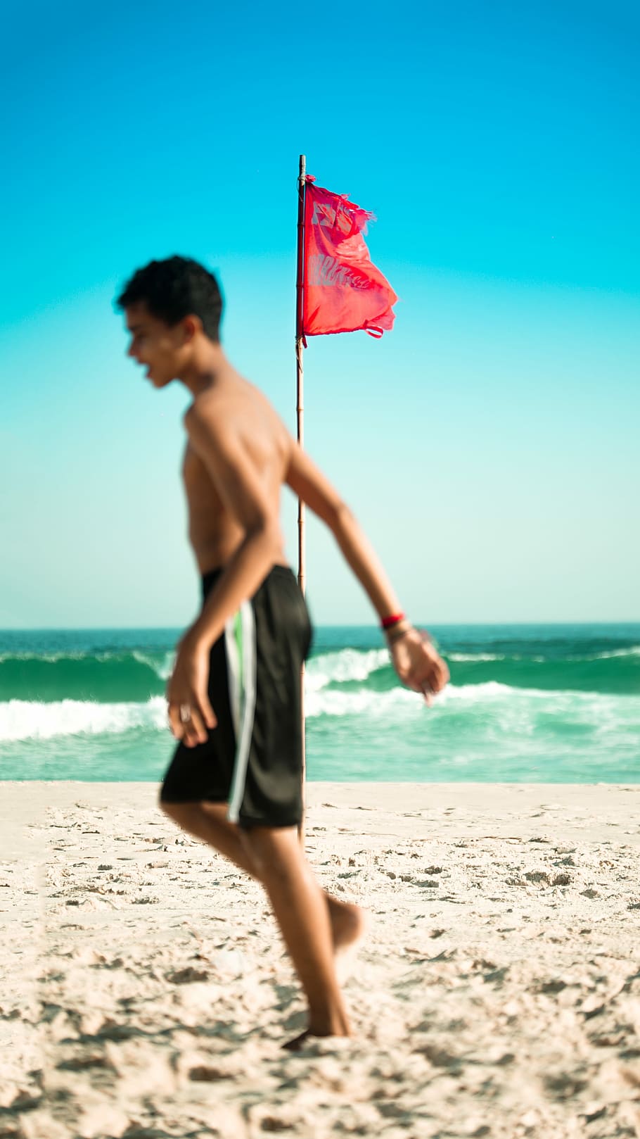 selective focus photo of red flag on brown pole near body of water, man in black shorts walking on seashore HD wallpaper