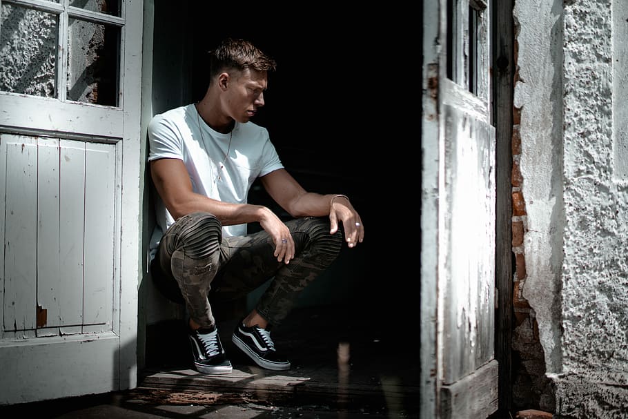 man in white crew-neck T-shirt sitting beside white door, man sitting beside gray wooden wall in front of opened door during daytime photo HD wallpaper