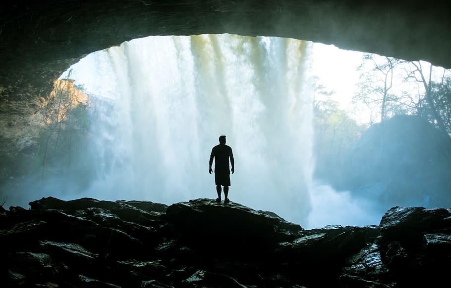 man standing on rock near cave, silhouette of man inside waterfall cave