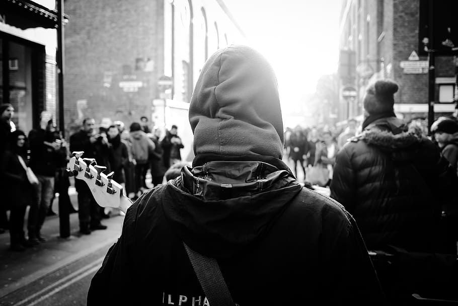 grayscale photography of person wearing hoodie, people gathered at street between buildings in grayscale photography
