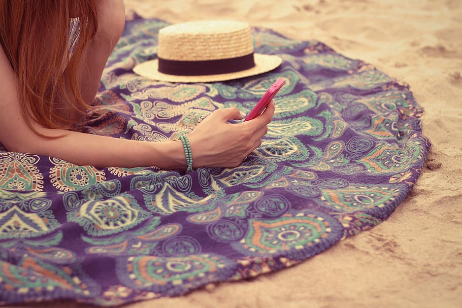 person holding red android phone and brown and black sun hat sitting in the white sand HD wallpaper