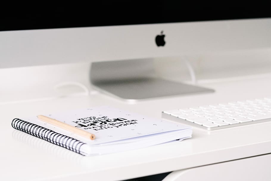 white book beside iMac and keyboard on table, brown pen on notebook beside keyboard HD wallpaper
