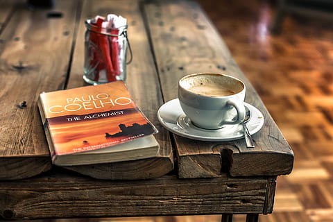 white and gray ceramic mug filled with coffee on white ceramic saucer with gray stainless steel spoon on brown wooden table, closeup photography of Paulo Coelho The Alchemist book near white ceramic teacup on saucer plate with stainless steel teaspoon on wooden table HD wallpaper