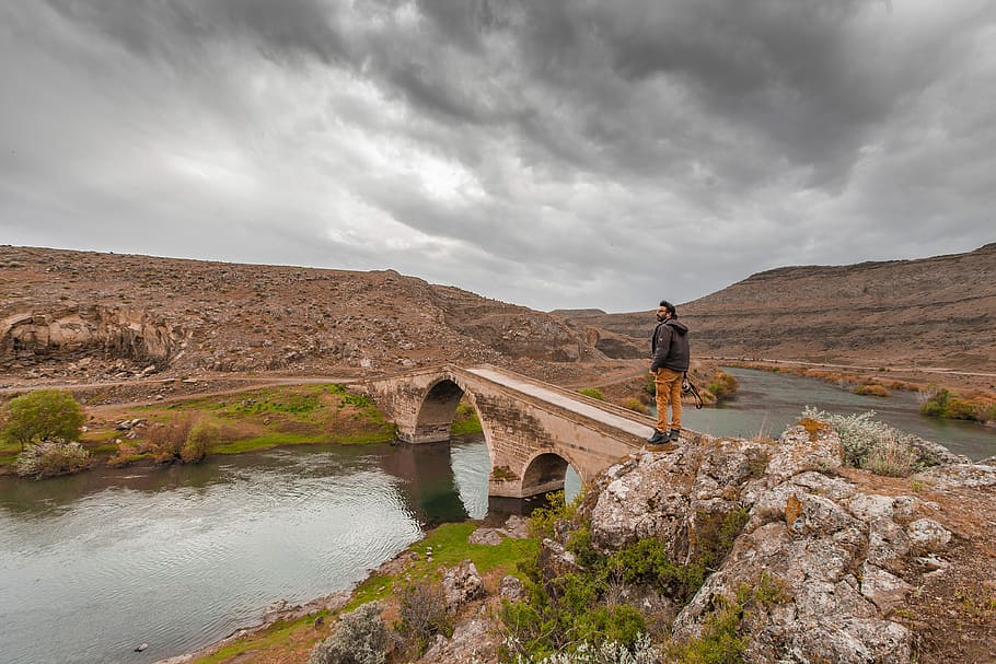 man standing on bridge of body of water, brown concrete bridge HD wallpaper