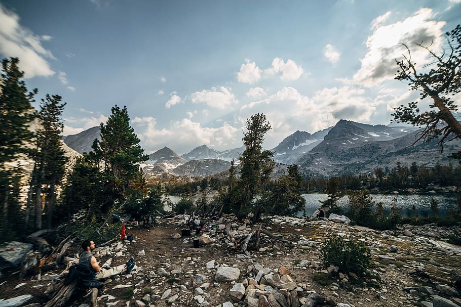 man sitting on ground in front of mountains and lake, man sitting on rock looking at river HD wallpaper
