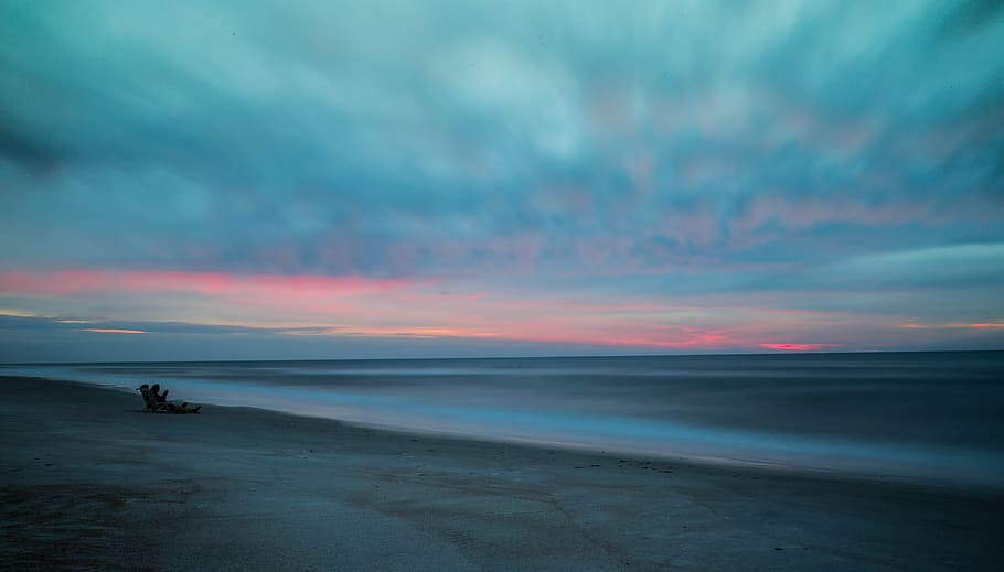 beach with drift wood, person sitting on gray sand near sea during dawn HD wallpaper